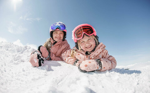 Zwei Kinder liegen lächelnd im Schnee und blicken in die Kamera.