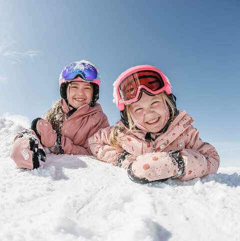 Zwei Kinder liegen lächelnd im Schnee und blicken in die Kamera.