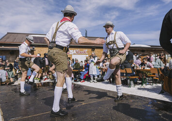 Männer in Lederhosen tanzen auf einer Bühne vor zahlreichem Publikum.