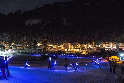Viele Menschen beim Nachtskifahren, mit leuchtenden Stöcken. Im Hintergrund die verschneiten und festlich beleuchteten Häuser.