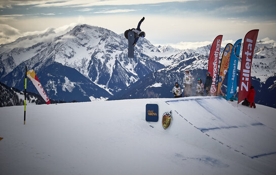 Snowboarder springt über eine Schanze mit Bergkulisse im Hintergrund.