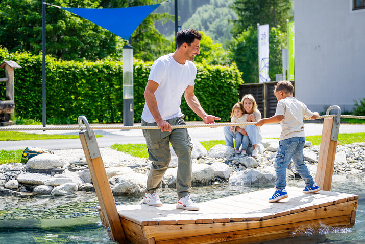 ein Vater und ein Sohn auf einem Floß, Mutter und Tochter schauen vom Ufer aus zu