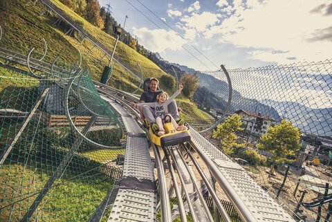 Ein Mann und ein Mädchen fahren mit einer Sommerrodelbahn.