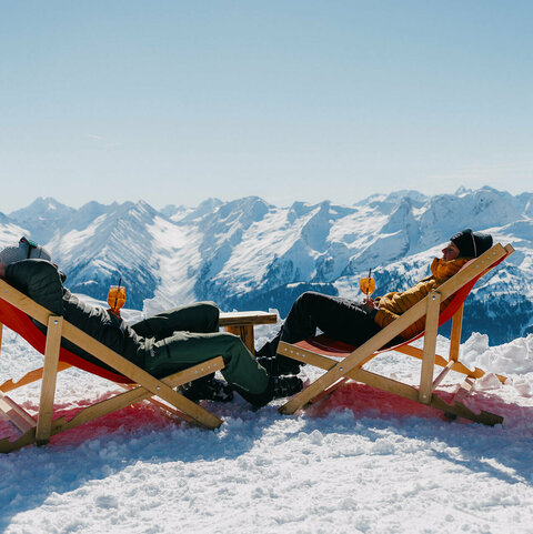 Zwei Personen entspannen auf Liegestühlen im Schnee vor einer verschneiten Berglandschaft.