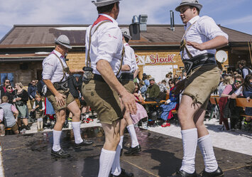 „Männer in Lederhosen tanzen auf einer Bühne vor Publikum.