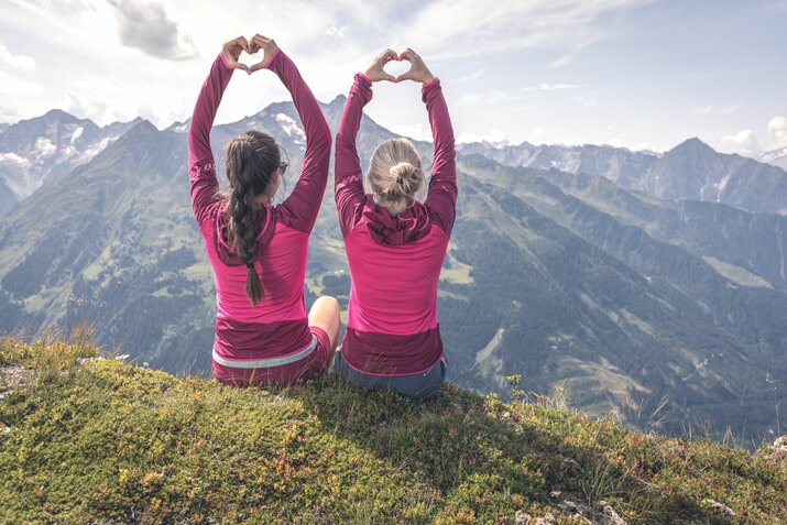 Zwei Frauen formen mit ihren Händen ein Herz und blicken auf eine atemberaubende Bergkulisse