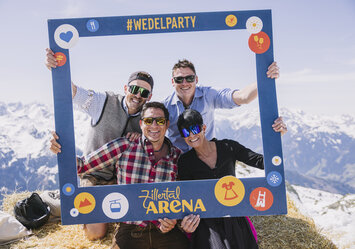 Drei Männer und eine Frau in Trachtenkleidung sitzen auf einem Heuballen und posieren mit einem Bilderrahmen.