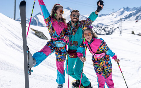 Drei fröhliche Skifahrer:innen in bunten Retro-Skianzügen posieren lachend im Schnee vor verschneiter Bergkulisse.