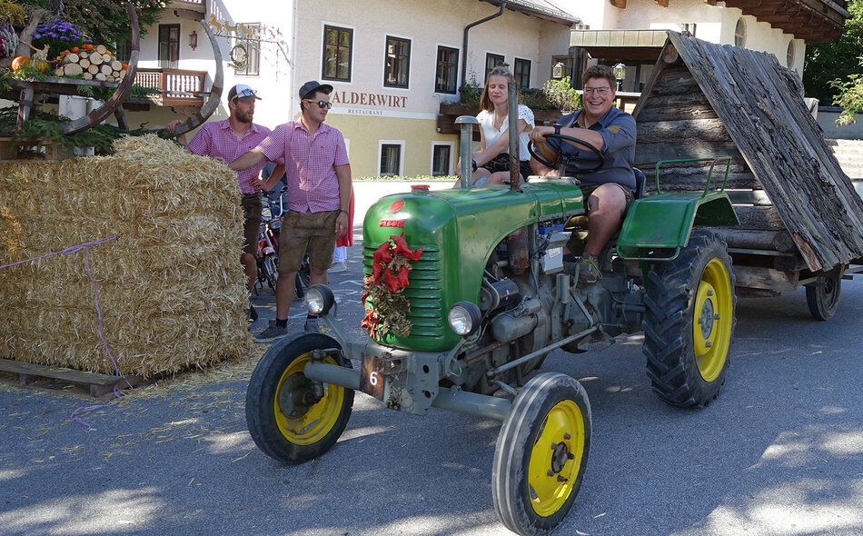 2 Leute sitzen auf Traktor mit angehaengter Holzhuette, daneben Zuschauer und Deko