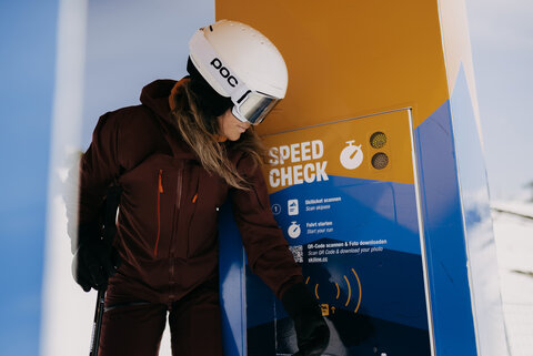 Eine Skifahrerin steht vor einem "Speed Check"-Terminal und scannt ihr Ticket, um ihre Geschwindigkeit zu messen.