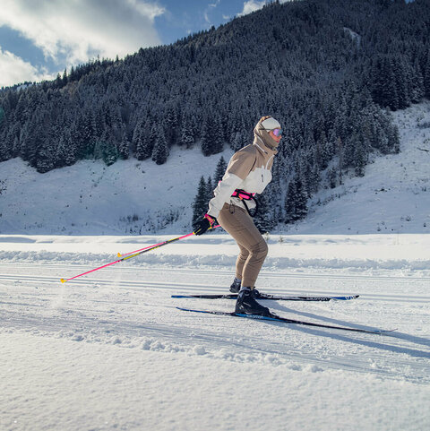 Eine Person fährt auf Langlaufskiern über eine verschneite Loipe vor einem bewaldeten Hang.