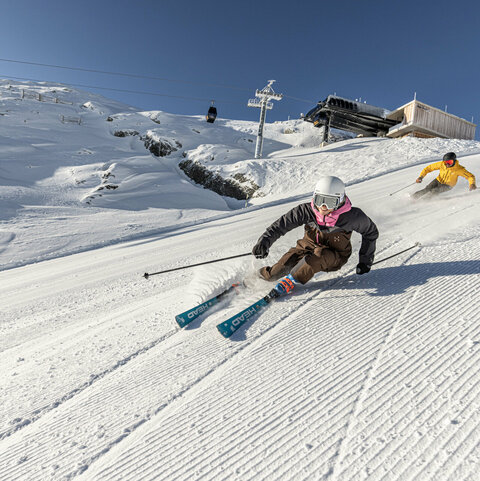 Zwei Skifahrer:innen fahren eine verschneite Piste hinab, im Hintergrund ein Sessellift und blauer Himmel.