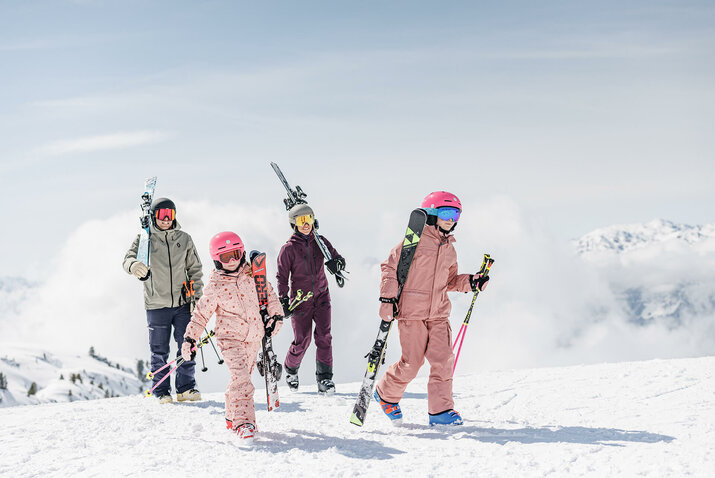 Zwei Erwachsene und zwei Kinder tragen Skier über eine verschneite Berglandschaft.Zwei Erwachsene und zwei Kinder tragen Skier über eine verschneite Berglandschaft.