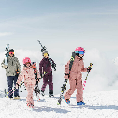 Zwei Erwachsene und zwei Kinder tragen Skier über eine verschneite Berglandschaft.Zwei Erwachsene und zwei Kinder tragen Skier über eine verschneite Berglandschaft.