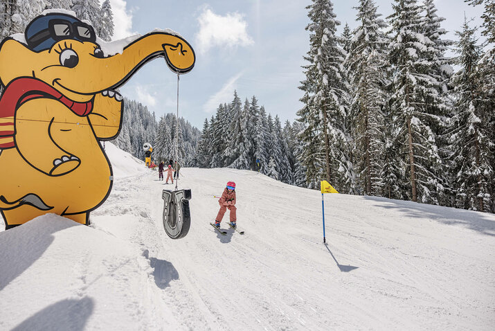 Kind fährt an einer gelben Elefantenfigur auf einer verschneiten Skipiste vorbei..