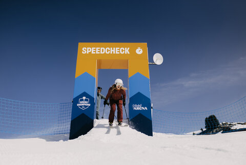 Skifahrerin in brauner Kleidung startet an einem gelb-blauen Torbogen mit der Aufschrift „SPEEDCHECK“ auf einer verschneiten Piste.