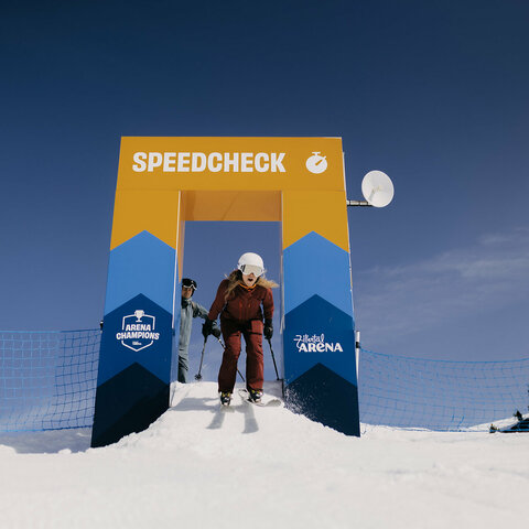 Skifahrerin in brauner Kleidung startet an einem gelb-blauen Torbogen mit der Aufschrift „SPEEDCHECK“ auf einer verschneiten Piste.