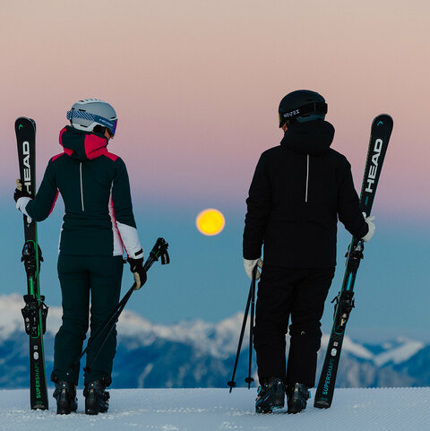 Zwei Skifahrer:innen stehen auf einer verschneiten Bergkuppe und schauen bei Dämmerung auf den aufgehenden Vollmond.