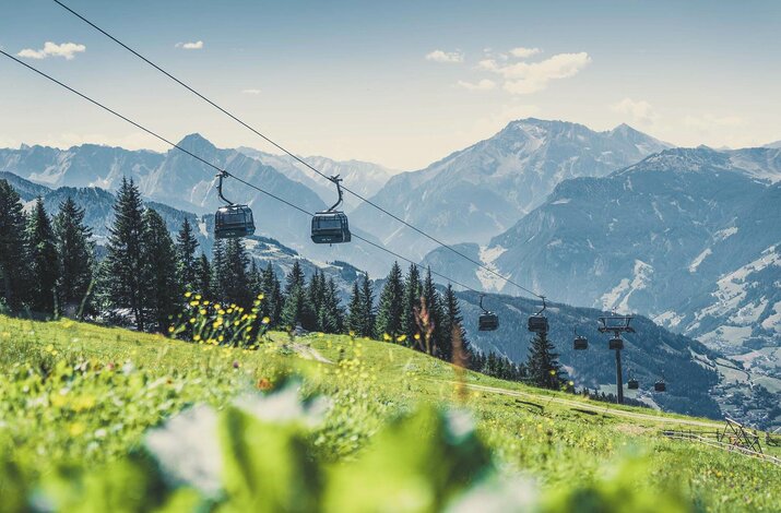 Sesselbahn fährt über eine grüne Almwiese mit Blick auf bewaldete Berghänge und ein Bergpanorama.