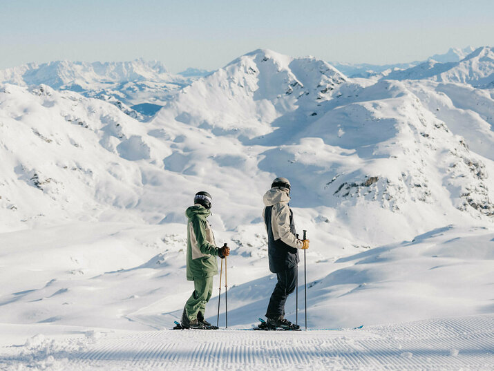 Zwei Skifahrer stehen auf einer präparierten Skipiste vor einer verschneiten Berglandschaft.