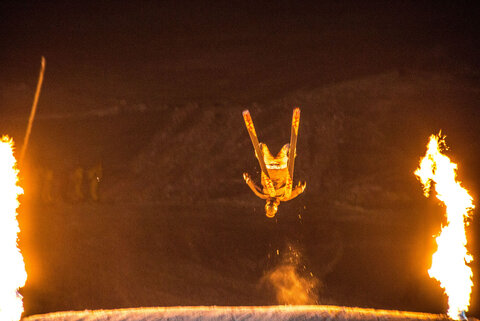 Ein Skifahrer macht einen Salto. Es ist Nacht und es brennen zwei Feuer.