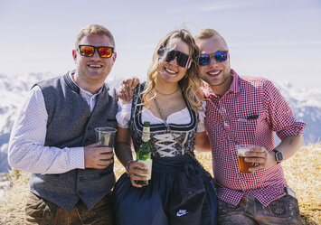 Zwei Männer und eine Frau in Tracht sitzen auf einem Heuballen.