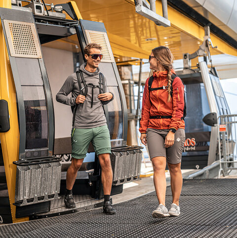 Zwei Wanderer steigen an der Bergstation aus einer Seilbahn aus.