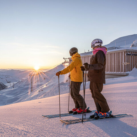 [Translate to EN:] Zwei Skifahrer:innen stehen auf einer verschneiten Piste und blicken bei Sonnenaufgang auf die umliegenden Berge.