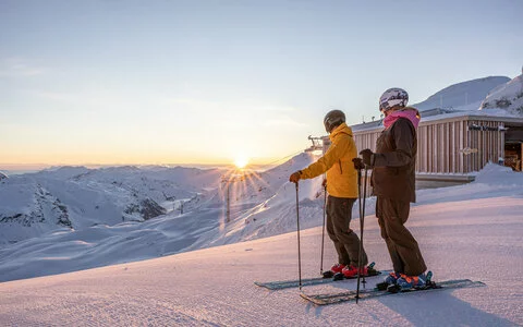 Zwei Skifahrer:innen stehen auf einer verschneiten Piste und blicken bei Sonnenaufgang auf die umliegenden Berge.