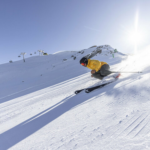 Ein Skifahrer in gelber Jacke fährt schnell eine sonnige, verschneite Piste hinab, mit Bergbahn und Bergen im Hintergrund.