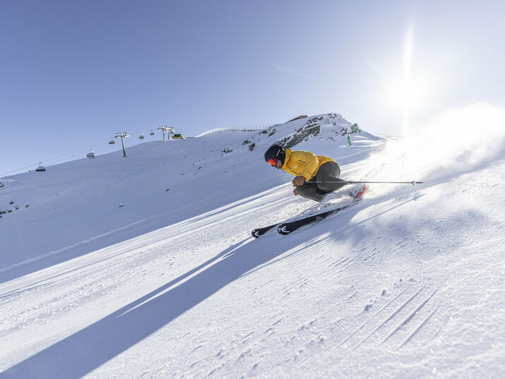 Ein Skifahrer in gelber Jacke fährt schnell eine sonnige, verschneite Piste hinab, mit Bergbahn und Bergen im Hintergrund.