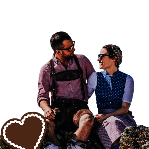 Ein Mann und eine Frau in Trachtenkleidung sitzen auf einem Stein, vor einer traumhaften Bergkulisse