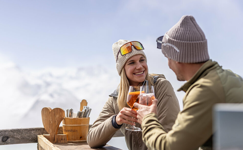 Ein Paar sitzt draußen an einem Holztisch in Bergkulisse und stößt mit Getränken an, beide lächeln.