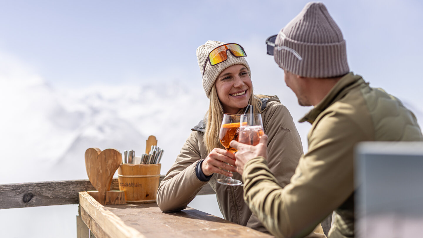 Ein Paar sitzt draußen an einem Holztisch in Bergkulisse und stößt mit Getränken an, beide lächeln.