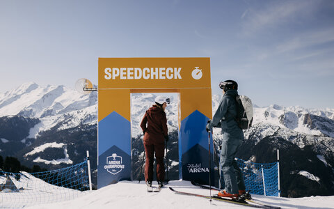 Zwei Skifahrer stehen vor einem Speedcheck-Tor auf einer schneebedeckten Bergpiste, im Hintergrund Berge und blauer Himmel.