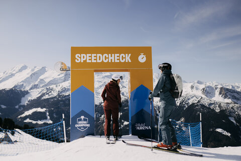 Zwei Skifahrer stehen vor einem Speedcheck-Tor auf einer schneebedeckten Bergpiste, im Hintergrund Berge und blauer Himmel.