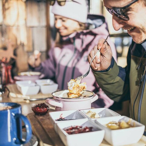 Ein Mann genießt Kaiserschmarrn auf der Sonnenterrasse einer Berghütte im Winter.