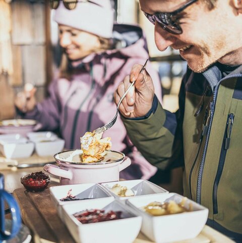 Ein Mann genießt Kaiserschmarrn auf der Sonnenterrasse einer Berghütte im Winter.