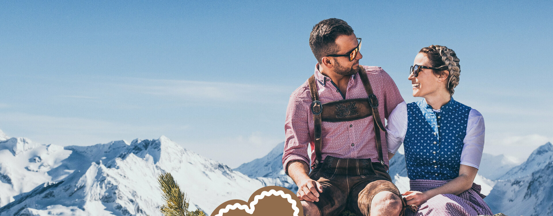 Ein Mann und eine Frau in Trachtenkleidung sitzen auf einem Stein, vor einer traumhaften Bergkulisse