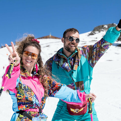 ChatGPT:  Zwei fröhliche Skifahrer in bunten retro Outfits posieren auf der Piste bei Sonnenschein.