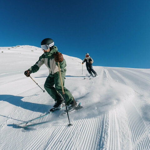 Zwei Skifahrer ziehen Schwünge auf einer firsch präparierten Skipiste unter blauem Himmel.