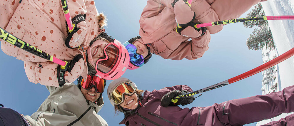 Vier Skifahrer blicken mit Skiern und Stöcken lächelnd von oben in die Kamera.