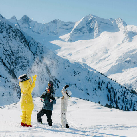 Das Maskotchen FUNty steht mit zwei Personen im Schnee vor einer verschneiten Berglandschaft.