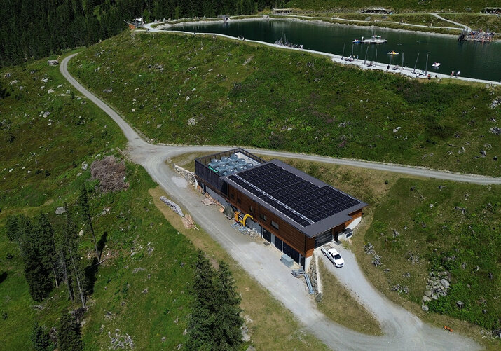 Luftaufnahme des Fichtensees mit Betriebsgebäude, Wanderwegen und umliegender Berglandschaft auf der Rosenalm.