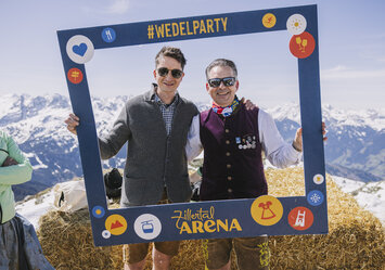 Zwei Männer in Trachtenkleidung posieren im Schnee mit einem Bilderrahmen.